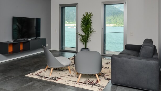 Un salon moderne doté d’un téléviseur à écran plat, de deux fauteuils gris, d’un canapé gris et d’une grande fenêtre offrant une vue sur un lac et des montagnes.