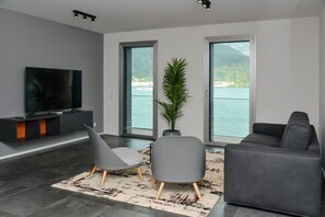 Een moderne woonkamer met een flatscreen televisie, twee grijze stoelen, een grijze bank en een groot raam met uitzicht op een meer en bergen.
