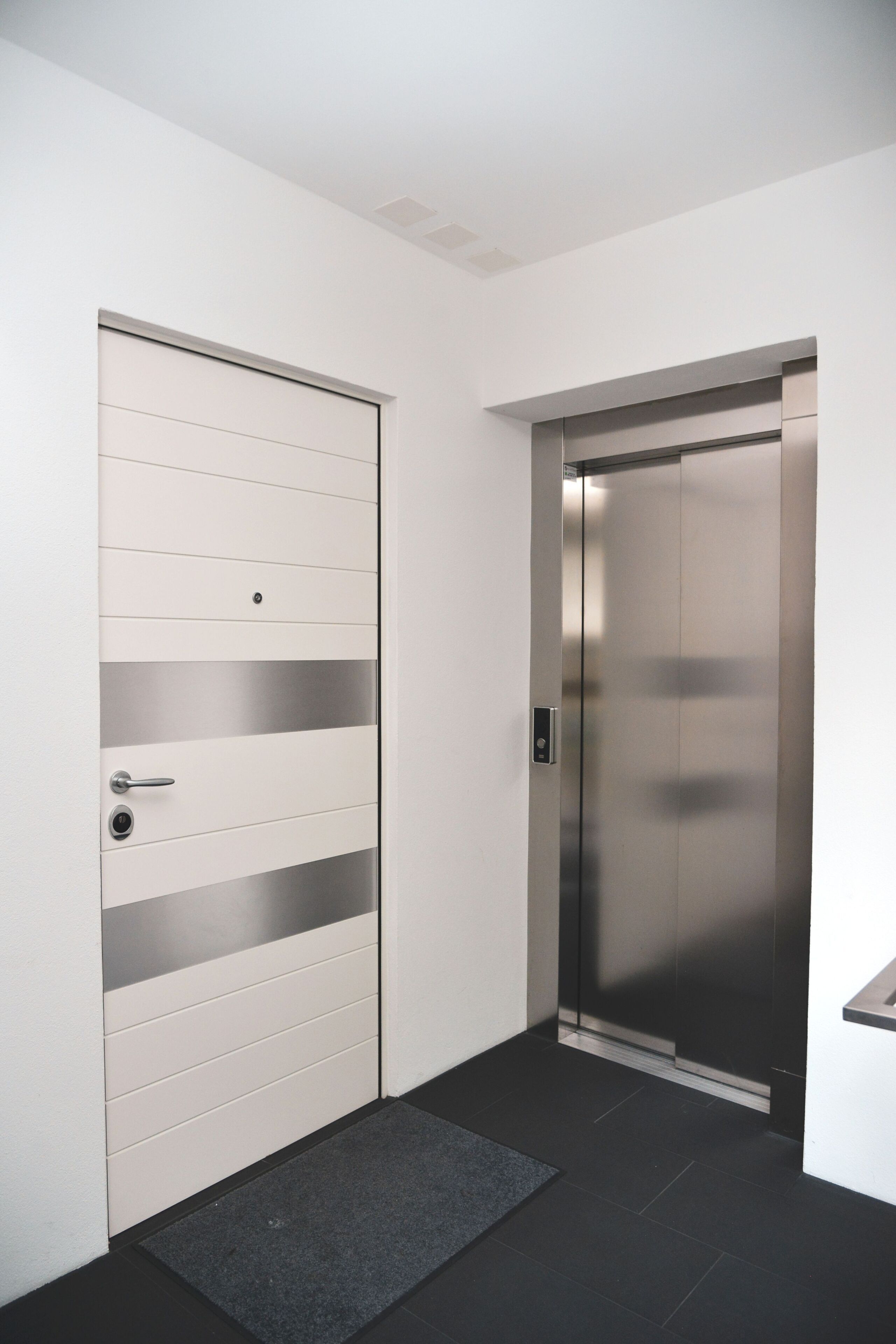 Een moderne lift en deur met een metalen afwerking, een klein kleedje bij de ingang, en een strak, minimalistisch design.