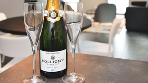 Une bouteille de champagne Colligny et deux verres sont posés sur une table dans un salon moderne, avec une table ronde et un canapé.