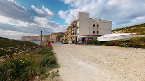 A coastal road with a building, a parked bus, and a boat. - Gozo Bus Glamping - Stay on a 1974 Vintage Maltese bus in Xlendi (Munxar)