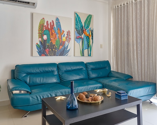 A teal leather sofa against a white wall with two colorful cactus paintings. A black coffee table with a blue vase and decorative items is in front.