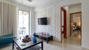A modern living room with a sofa, a coffee table with decorative items, a TV on a white wall, and a view of the outside through a window. - Apto En Zona Privilegiada De Santo Domingo City (Santo Domingo)