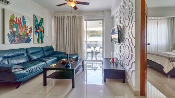 A modern living room with a blue sofa, a coffee table, and a ceiling fan. There is a balcony with a view of the outside. - Apto En Zona Privilegiada De Santo Domingo City (Santo Domingo)
