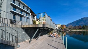 Um edifício moderno com varanda com vista para um lago, um deck de madeira e uma visão clara da água e montanhas ao fundo.