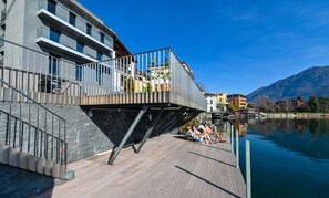A modern building with a balcony overlooking a body of water, a wooden deck, and a mountain in the background.