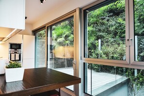 A modern kitchen with a built-in oven, a dining area with a wooden table, and large sliding glass doors leading to an outdoor deck with greenery.