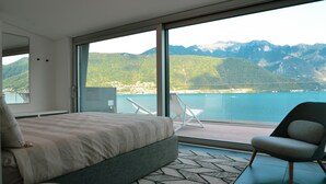 Quarto moderno com uma cama grande, área de estar com uma poltrona e vista para montanhas e água através de amplas janelas.