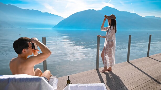 Um casal em um deck de madeira com vista para um lago calmo e montanhas ao fundo.