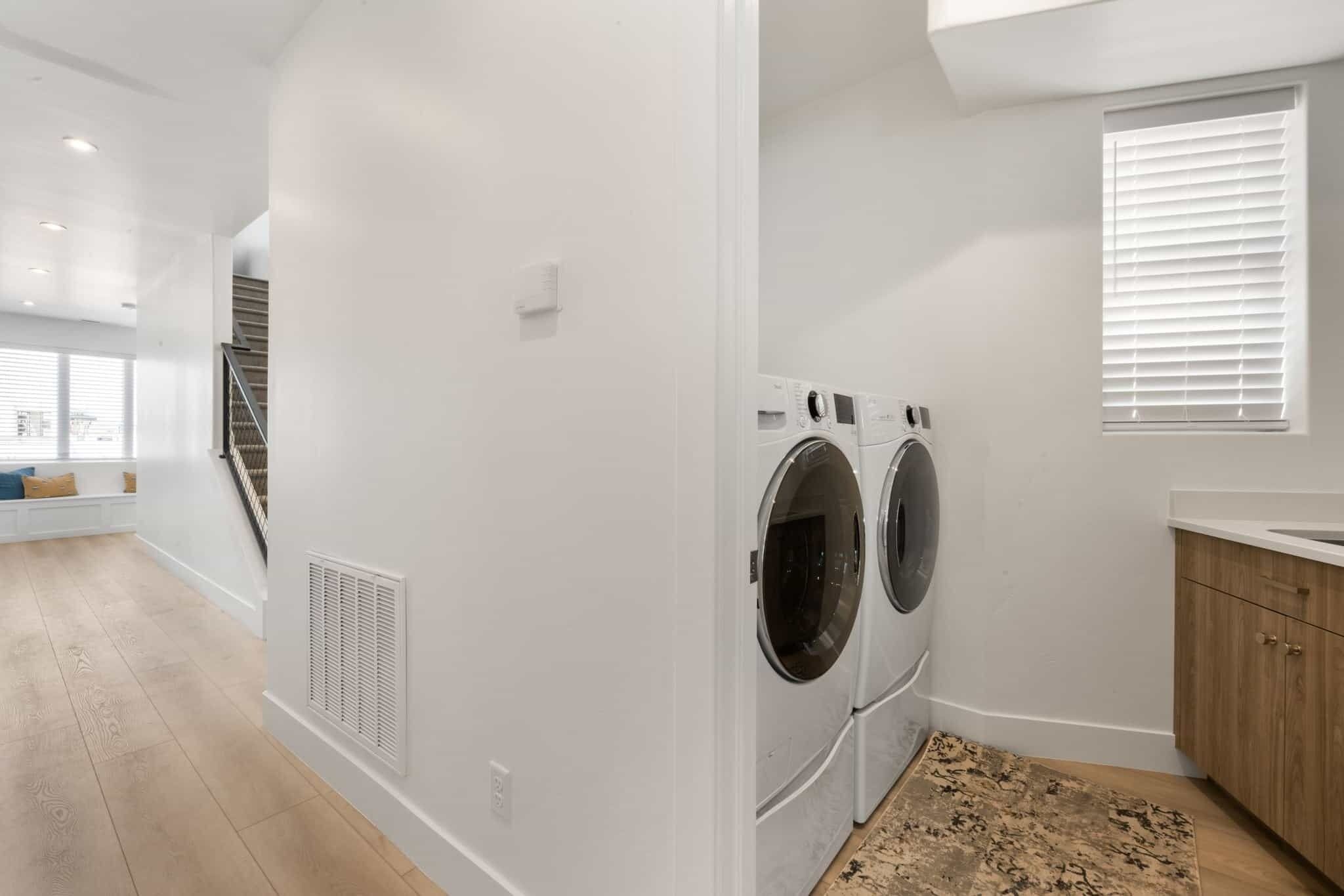 A laundry room with two front-loading washers and dryers, a wooden floor, and a window with blinds.