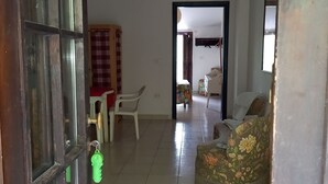 A spacious living room with a tiled floor, a floral sofa, and a white chair. A wooden door with a keyhole is visible on the left.