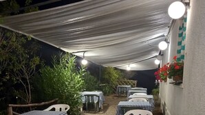 An outdoor patio area with tables and chairs, covered by a white canopy, and illuminated by hanging lights.