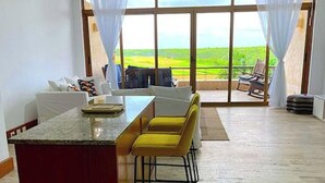 A spacious living area with a kitchen island, yellow bar stools, and a large sliding glass door leading to a balcony with a view of a green landscape. - Srvittinivillas Aptla/p11 Great Apartment /work Travel / Couple /altos de Chavon (La Romana)