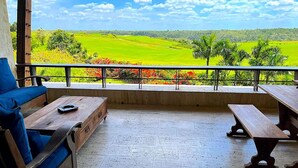 A balcony with a wooden table and a blue sofa, overlooking a green landscape with fields and trees. - Srvittinivillas Aptla/p11 Great Apartment /work Travel / Couple /altos de Chavon (La Romana)