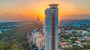 A high-rise building with a unique rooftop design, surrounded by a dense urban area with various other buildings and a clear view of the sunset. - Srvittinivillas Aptla/p11 Great Apartment /work Travel / Couple /altos de Chavon (La Romana)