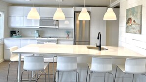 Uma cozinha moderna com armários brancos, uma ilha central com banquetas, iluminação pendente, eletrodomésticos de aço inoxidável e uma pintura na parede.