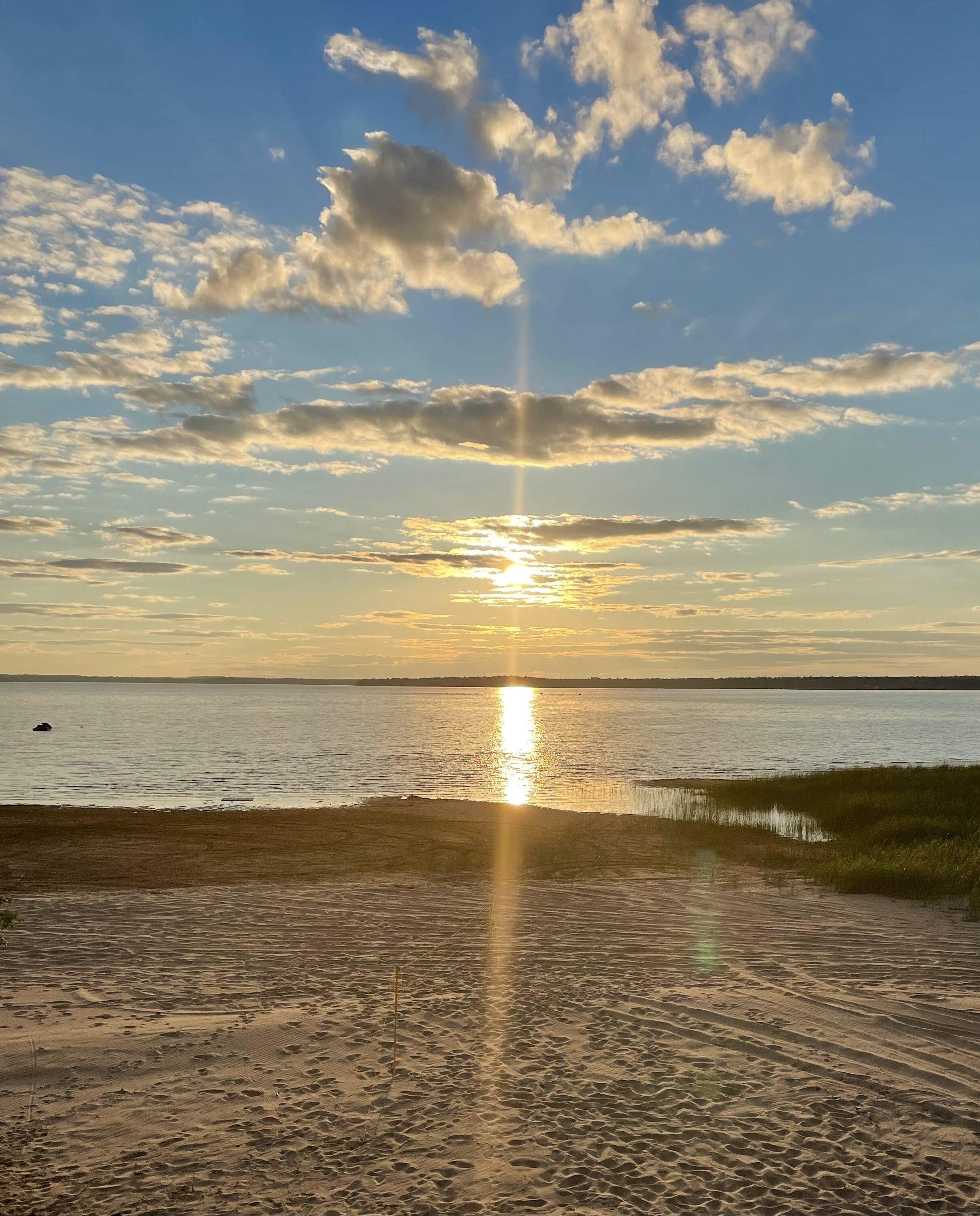 A beach with a sunset over the water, a sandy path leading to the water, and a distant shoreline.