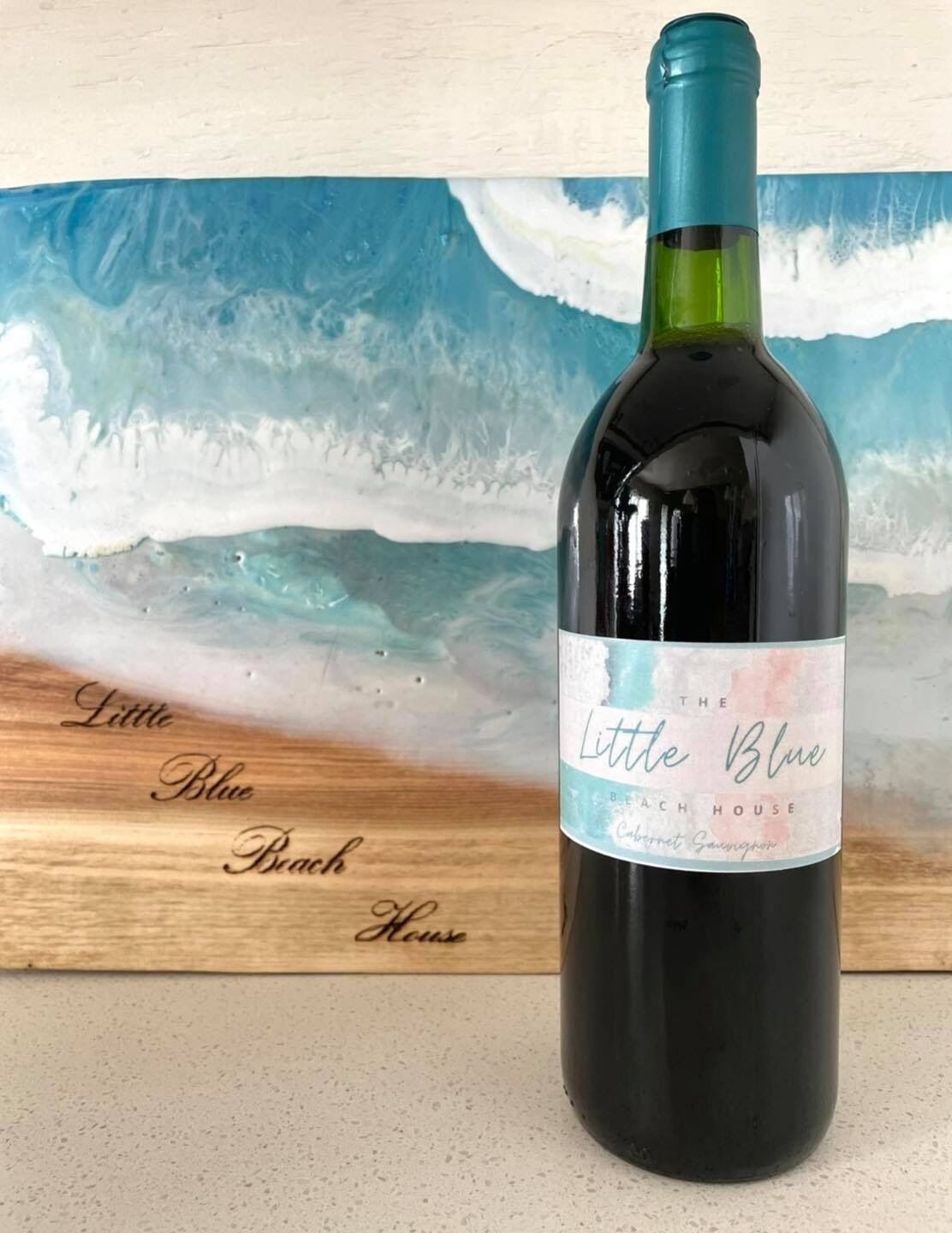 Una botella de vino con la etiqueta "The Little Blue Beach House Cabernet Sauvignon" colocada frente a una tabla de madera con el mismo nombre.