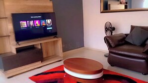 A living room with a flat-screen TV on a wooden stand, a brown leather sofa, and a round wooden coffee table. - Enjoy This Fully Furnished Apartment in Chililabombwe (Chililabombwe)