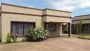 A building with a beige exterior, multiple windows, and a paved entrance area with a small garden. - Enjoy This Fully Furnished Apartment in Chililabombwe (Chililabombwe)