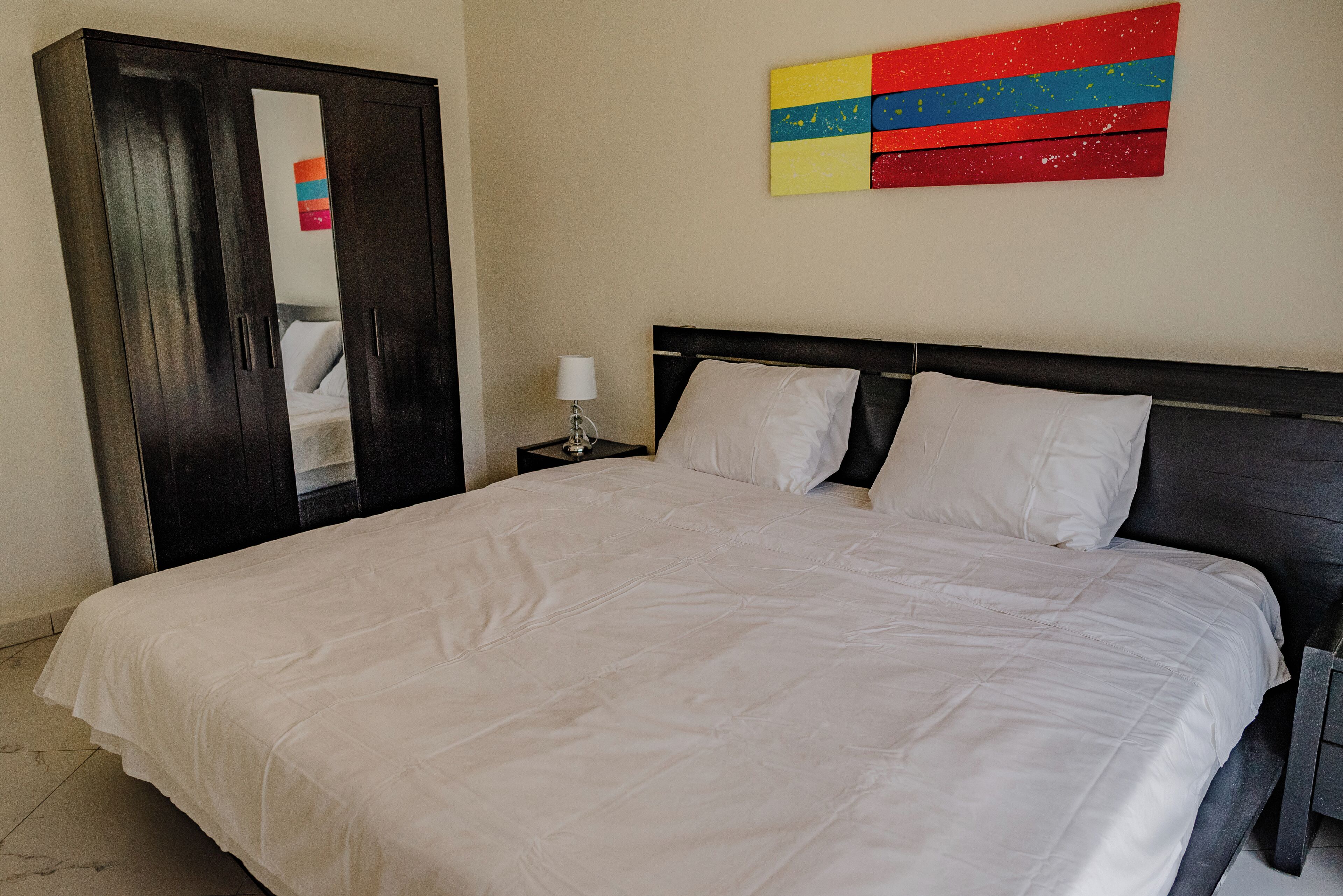 A neatly made bed with white bedding, a dark wooden wardrobe, and a nightstand with a lamp.