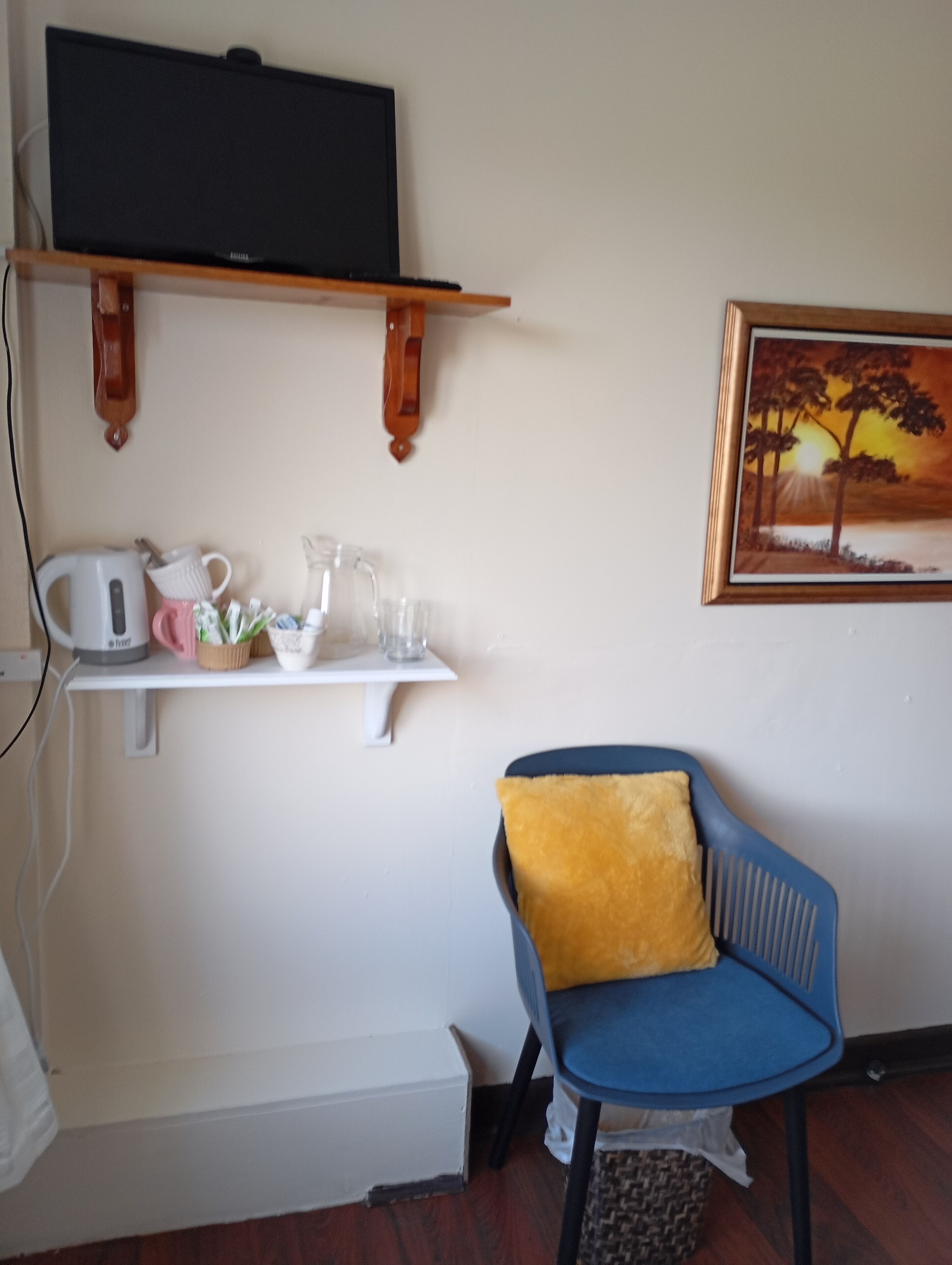 A room with a television on a wooden shelf, a white shelf with a kettle, a pink mug, and a glass pitcher. There's a blue chair with a yellow cushion and a framed painting of a sunset on the wall.