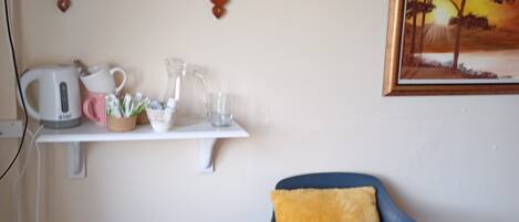 A room with a television on a wooden shelf, a white shelf with a kettle, a pink mug, and a glass pitcher. There's a blue chair with a yellow cushion and a framed painting of a sunset on the wall.