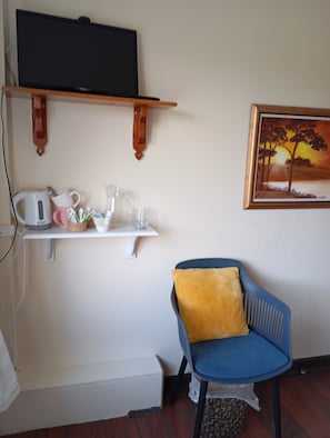 A room with a television on a wooden shelf, a white shelf with a kettle, a pink mug, and a glass pitcher. There's a blue chair with a yellow cushion and a framed painting of a sunset on the wall.