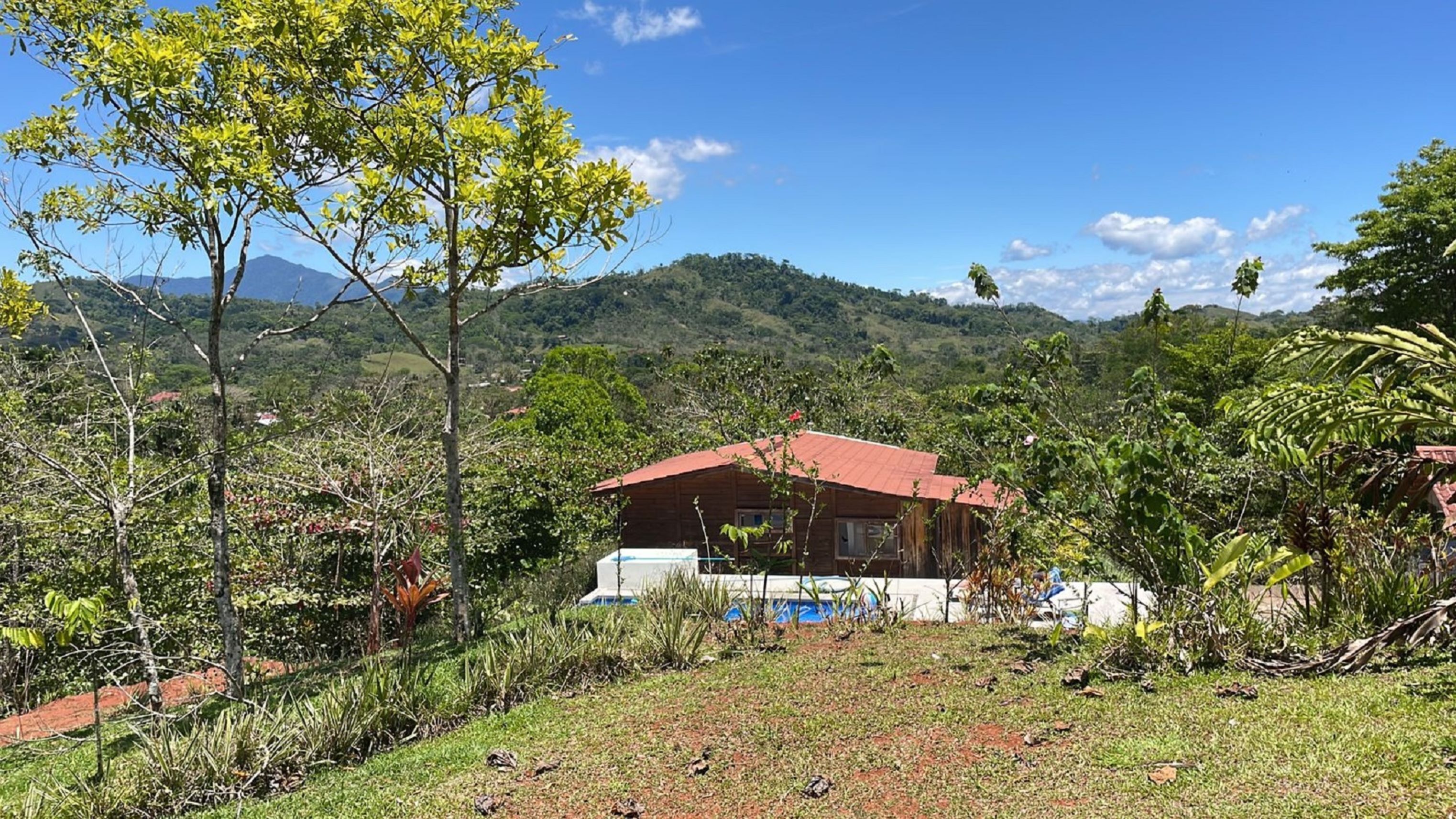 Une cabane en bois avec un toit rouge, entourée d’une végétation luxuriante et avec des collines en arrière-plan.