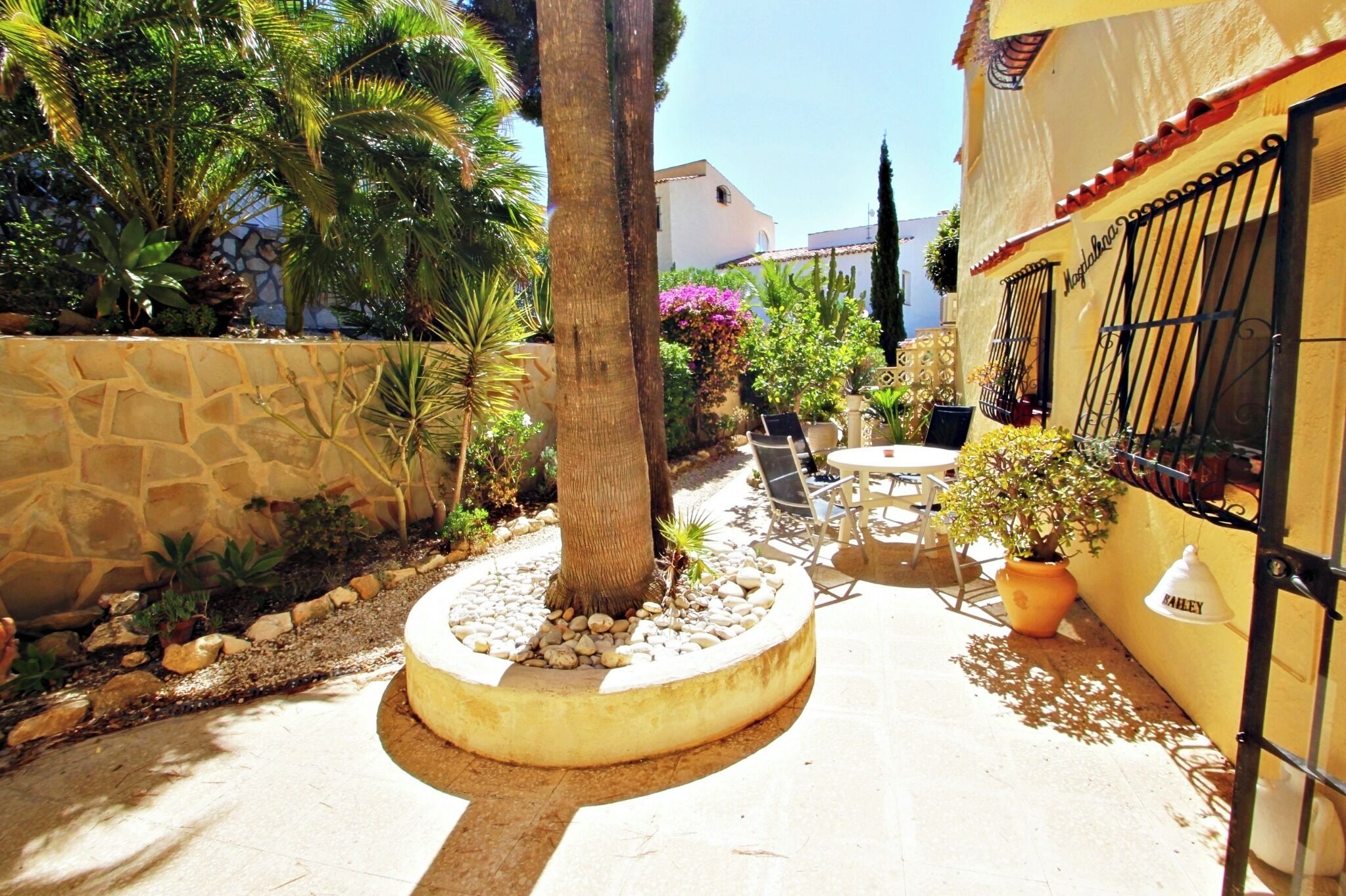 A courtyard with a stone wall, palm trees, and a seating area with chairs and a table.