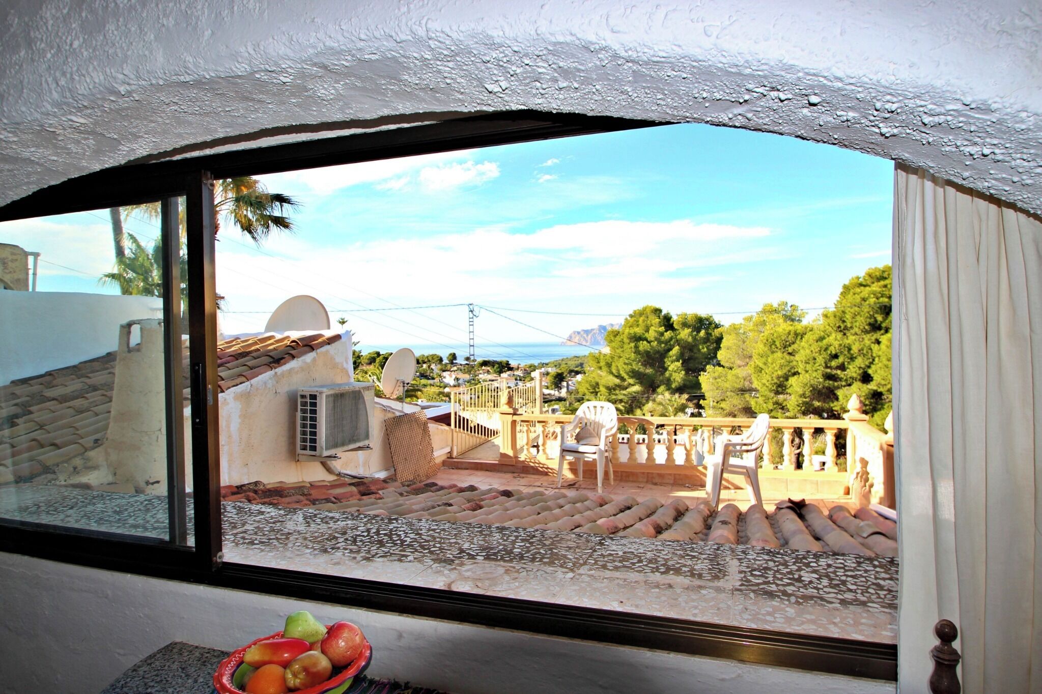 Blick aus einem Fenster auf eine Terrasse mit Steindeckung, Gartenmöbeln und eine ferne Landschaft mit Bäumen und Hügeln.