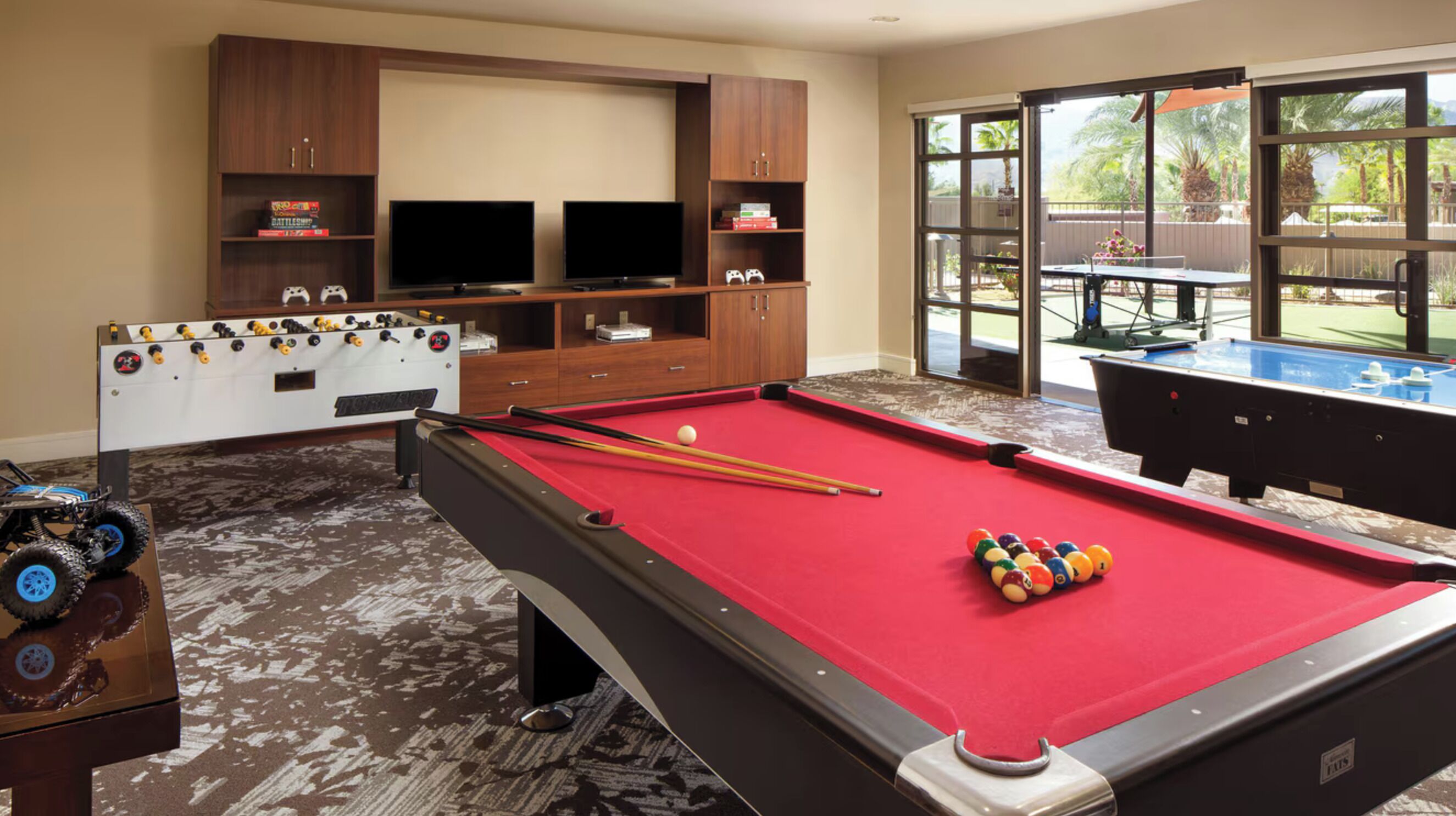 A pool table with a red felt surface, a foosball table, and a television set on a wooden cabinet.