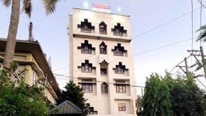 Um edifício de vários andares com telhado plano e uma placa que indica ser um hotel. Há várias janelas em cada andar, e o edifício é cercado por vegetação.