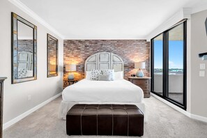 A bedroom with a brick wall, a large bed, two bedside lamps, a bench, and a view through a sliding door. - Destin Harbor & Crab Island Views ~ Updated 3 Bedroom Condo! (Destin)