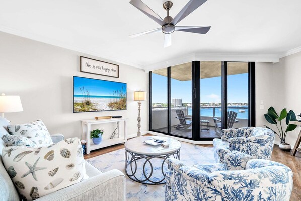 A modern living room with a ceiling fan, a large window with a view of the sea, a painting, and a dining area with outdoor seating. - Destin Harbor & Crab Island Views ~ Updated 3 Bedroom Condo! (Destin)