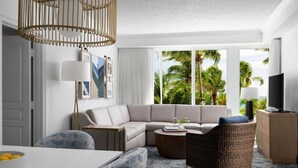 Un moderno salón con sofá, sillones, una mesa de centro y televisor. Amplias ventanas ofrecen vistas a palmeras y al océano.