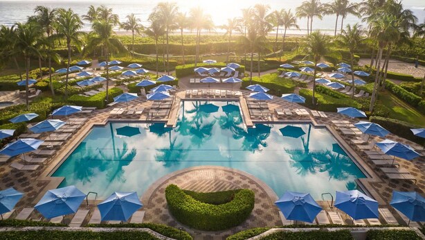 Zona de piscina en un resort con múltiples tumbonas y sombrillas, rodeada de palmeras y vista al océano.