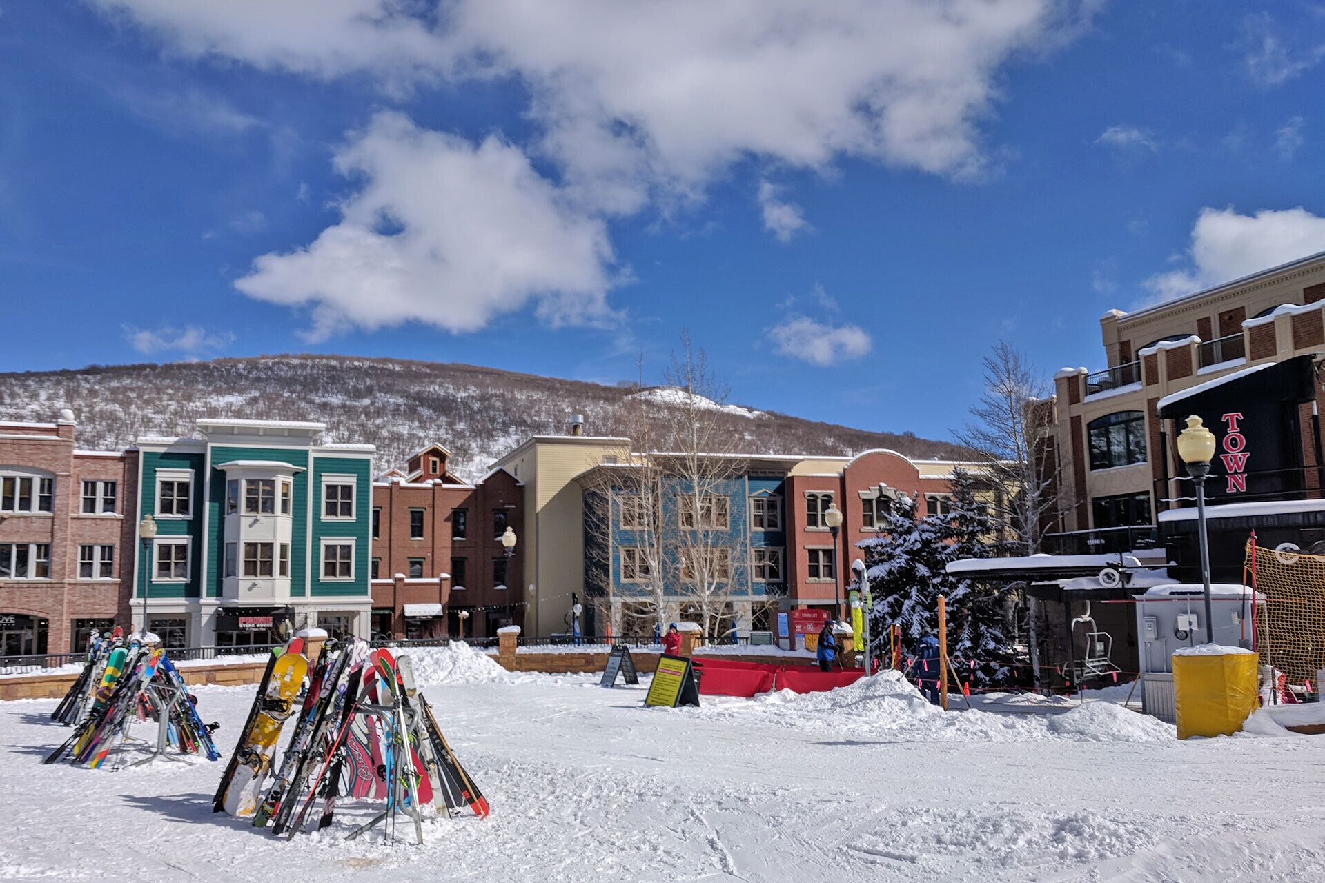 A snow-covered outdoor area with a row of colorful buildings, a ski rack, and a signboard.