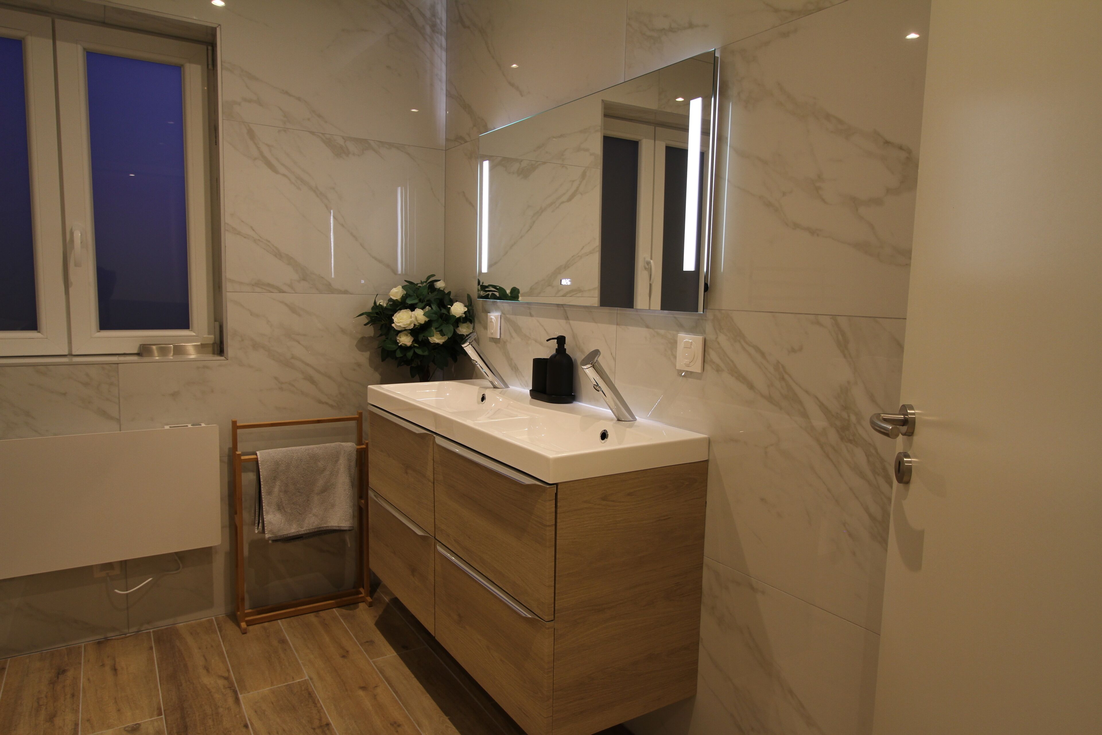 A modern bathroom with a marble floor, a wooden vanity with a mirror, and a window with frosted glass.