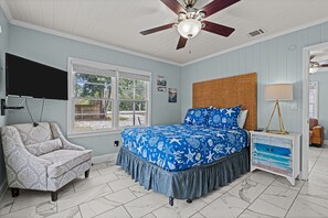 A bedroom with a bed, a chair, a TV, a ceiling fan, and a window. - Coastal Cabana ~ Cozy getaway in the heart of town w/a privately fenced backyard (Panama City Beach)