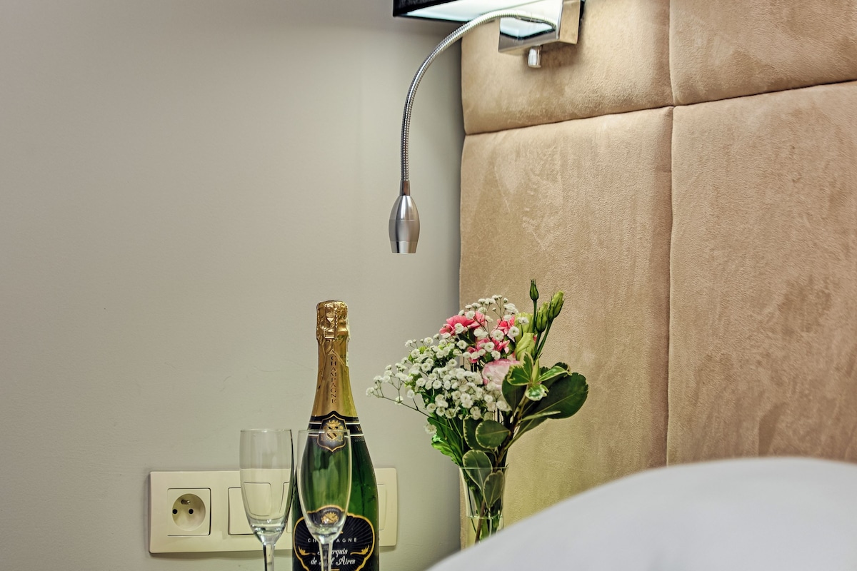 A hotel room with a bed, a nightstand with a bottle of champagne and two glasses, a vase with flowers, and a wall-mounted lamp.