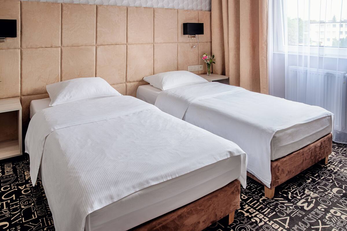 A hotel room with two beds, a TV mounted on the wall, a nightstand with a vase of flowers, and a window with curtains.