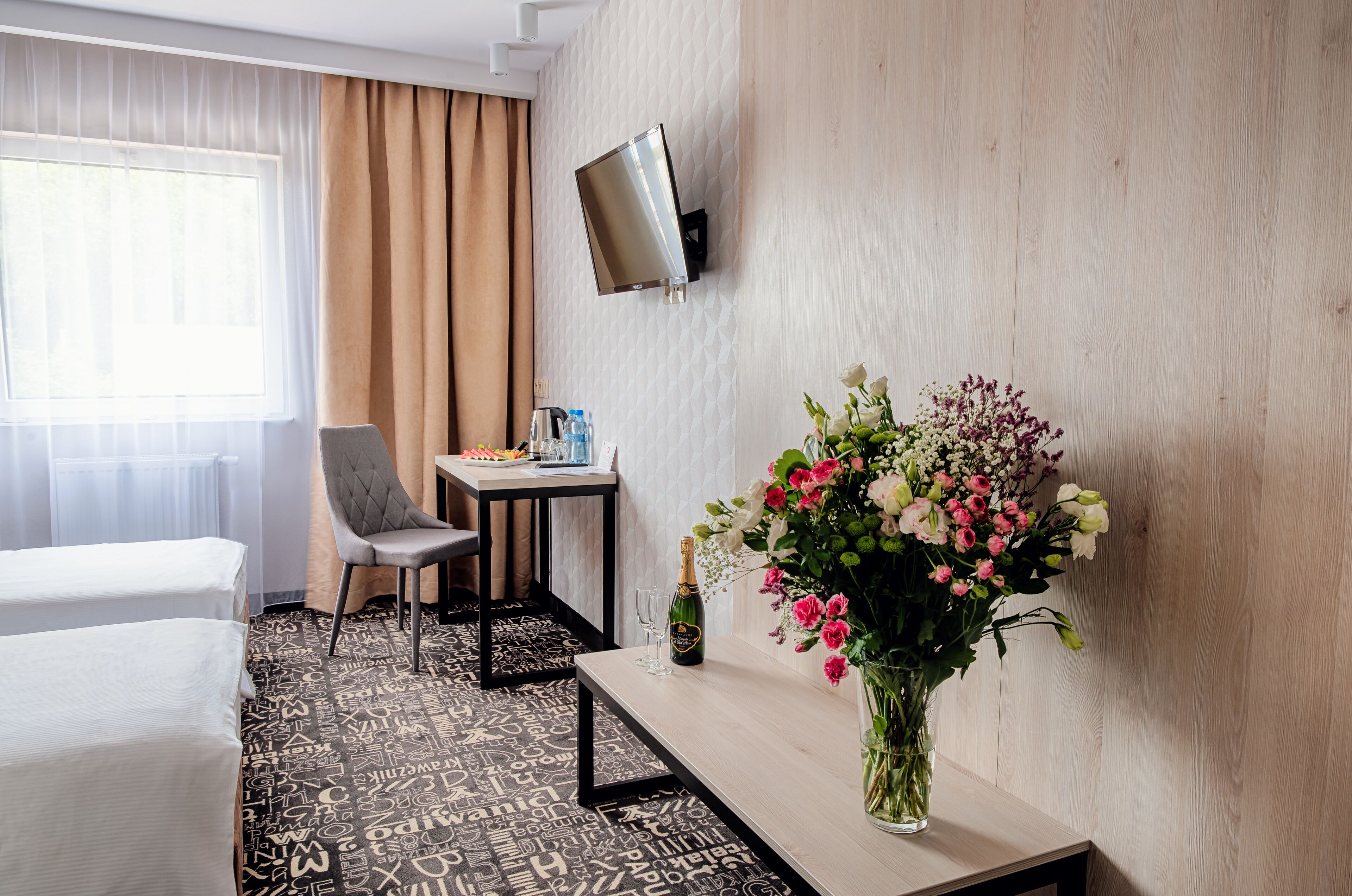 Habitación de hotel con una cama, una silla, una mesita con una botella y vasos, un televisor montado en la pared y un arreglo floral sobre la mesita.
