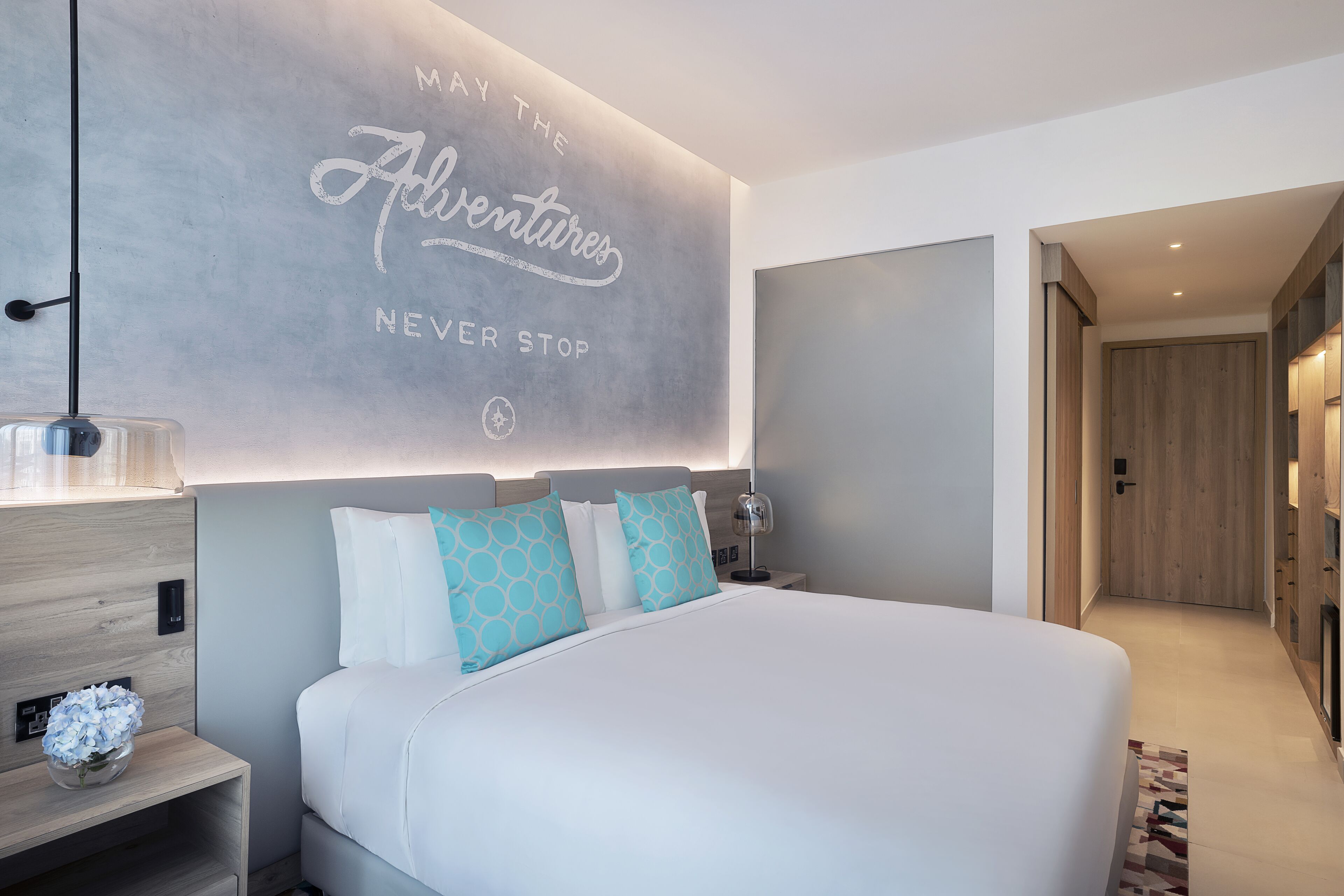 Une chambre d’hôtel moderne avec un grand lit, une table de chevet ornée d’un vase de fleurs, un mur sur lequel figure un texte inspirant, et une porte en bois.