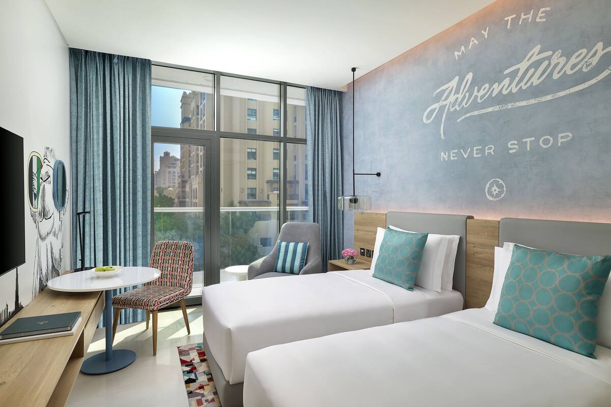 A hotel room with two beds, a desk, a chair, and a large window with curtains.