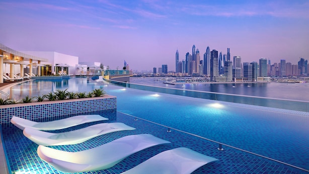 A modern infinity pool with a city skyline view, lounge chairs, and a waterfront promenade. - NH Collection Dubai The Palm (Dubai)