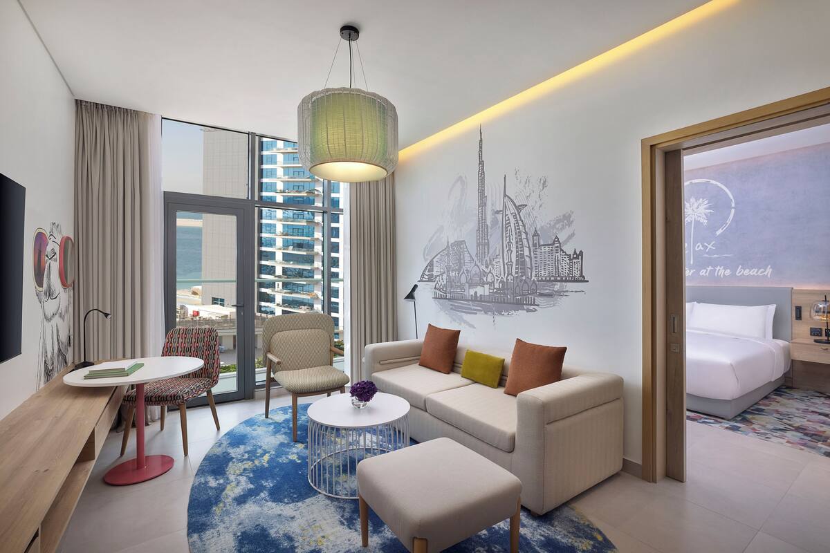 A modern hotel room with a large window, a sofa, a coffee table, and a bed.