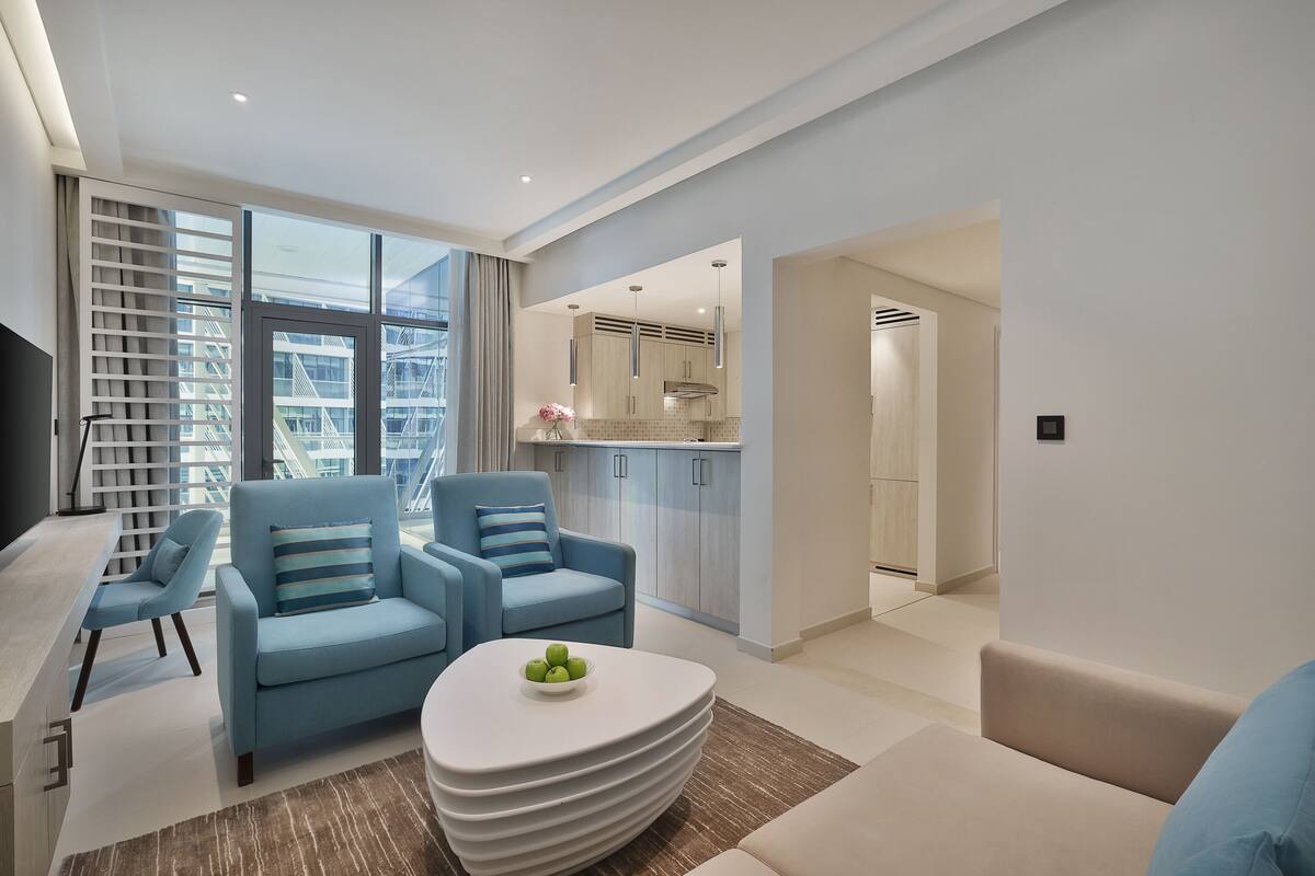 A modern living room with a sofa, armchairs, and a coffee table. There is a kitchen area visible through an open door.
