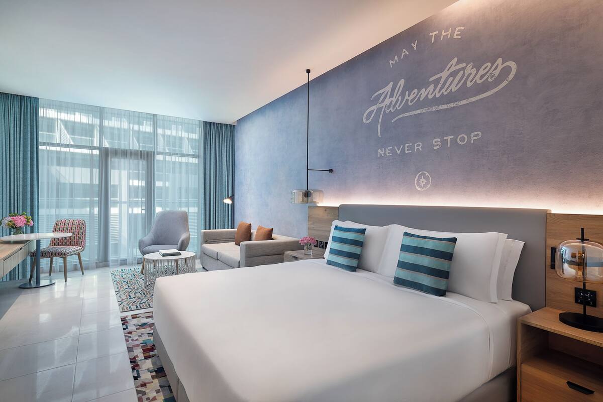 A modern hotel room with a large bed, a sofa, a desk, and a chair. There is a large window with curtains, a wall with a motivational quote, and a small table with a vase of flowers.