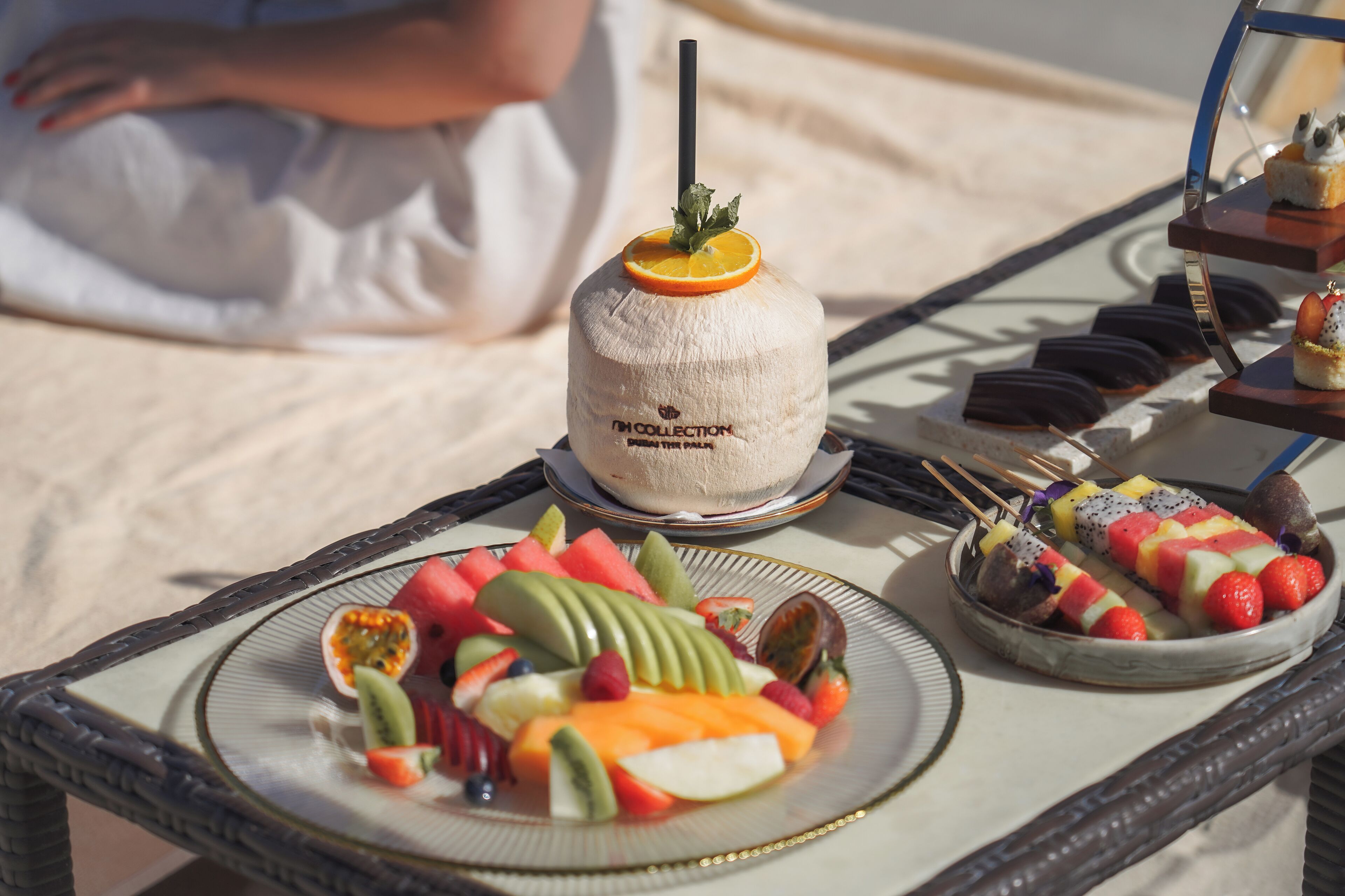 Eine tropisch gestaltete Dessert-Anrichtung mit Kokosgetränk, Obstspießen und verschiedenen Gebäcken auf einem Korbtisch.